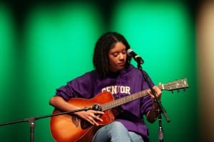young woman in a purple hoodie sitting onstage with a guitar